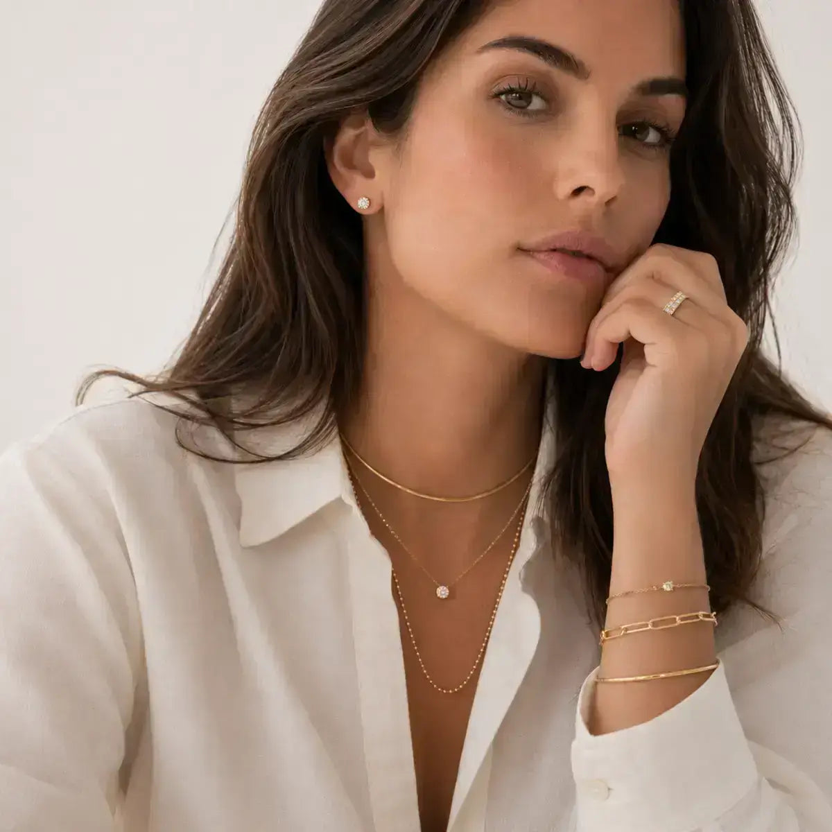 Mulher elegante a usar colares dourados em camadas, pulseiras, anel e brincos minimalistas com camisa branca em fundo neutro, destacando joalharia feminina sofisticada, acessórios modernos e estilo intemporal de luxo.