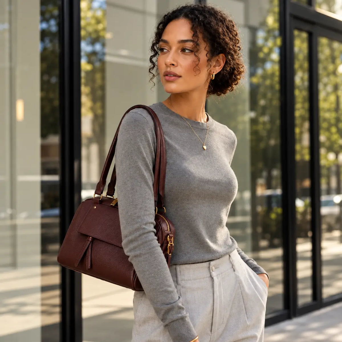 Modelo elegante com mala castanha de design sofisticado, camisola cinzenta e calças bege, num cenário urbano moderno, com acessórios dourados e estilo minimalista.
