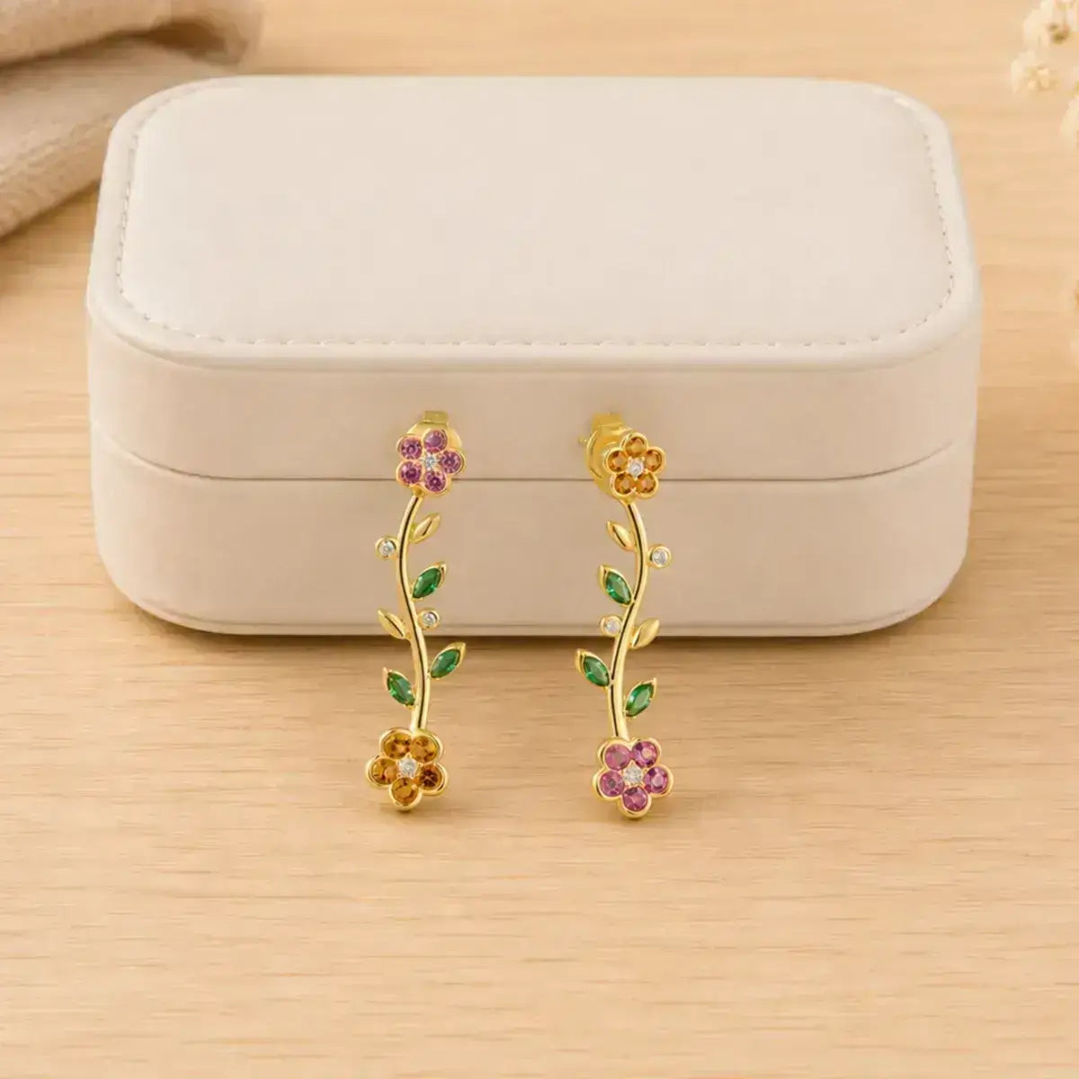 Brincos compridos design floral em prata 925 dourada e pedras coloridas brilhantes encostados numa caixa de joias branca